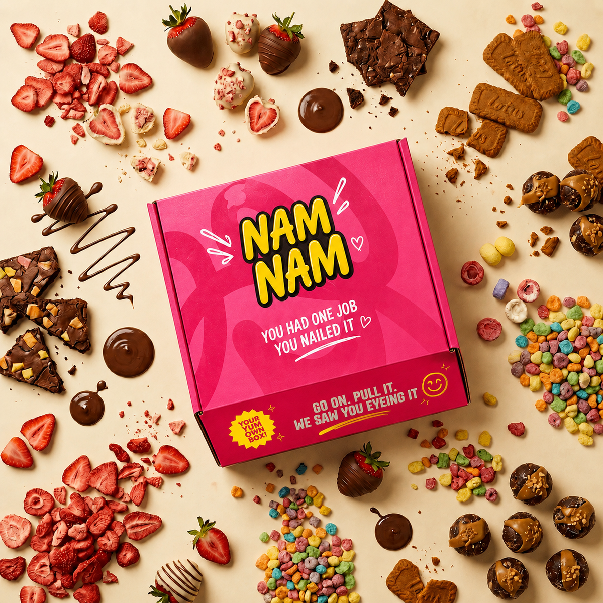 A bright pink rectangular box with bold yellow 'NAM NAM' lettering sits on a beige surface surrounded by assorted chocolate-covered strawberries, candy corn, pretzels, cookies, and candy pieces. The box has a humorous slogan printed on the front.